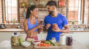 Un couple d'athlètes en tenue de sport dans une cuisine accueillante, la femme tenant un verre de boisson foncée, tandis que l'homme tient un pot de légumes fermentés et des légumes frais sur la table devant eux.
immunicure.com